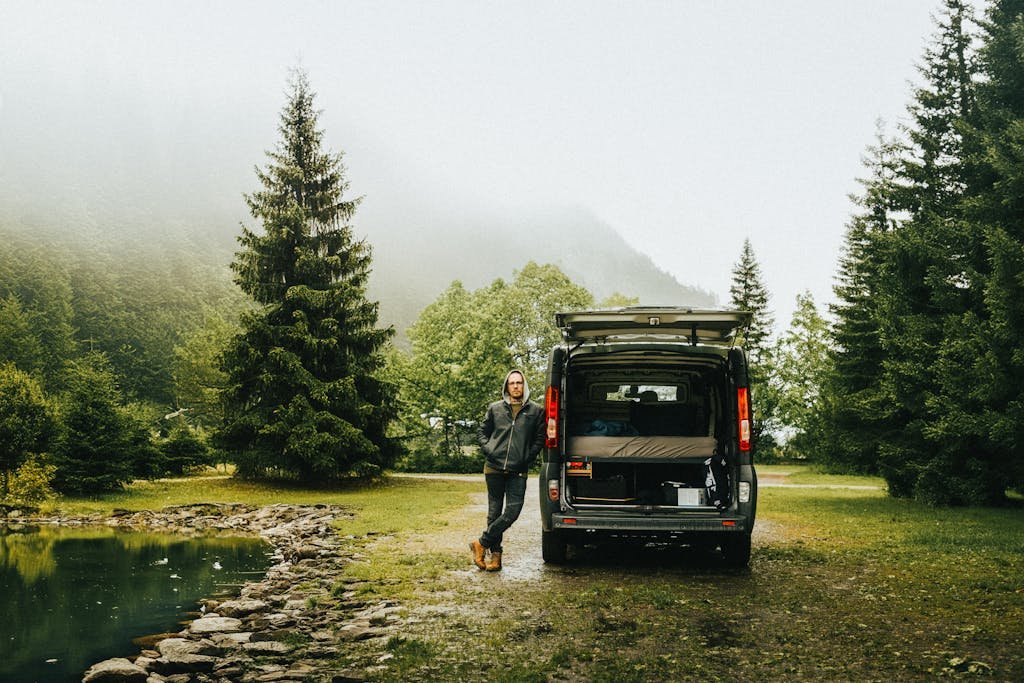 A man leans against an open van in a misty mountain park with conifer trees.